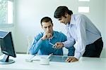 Two young businessmen in office, using cell phone, looking down