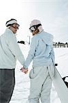 Two young friends walking, holding hands, looking over shoulders, one carrying snowboard, rear view