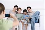 Young girl taking photo of her parents and brothers sitting together on sofa