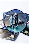 Miniature businessmen shaking hands on pile of credit cards, viewed through magnifying glass