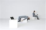 Identical businessmen, one using laptop while the other lies on back, exhausted