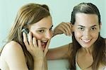 Two young friends smiling together, using cell phone, one looking at camera