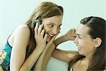 Two young friends smiling at each other, one using cell phone