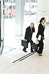 Business associates walking in lobby, one using cell phone, full length