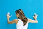 Woman standing with hands against blue wall, looking over shoulder, rear view