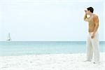 Man standing at the beach, looking at sailboat in the distance, side view