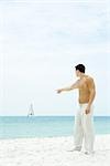 Man standing at the beach, pointing at sailboat in the distance, side view