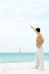 Man standing at the beach with arm raised, waving at sailboat in the distance, side view