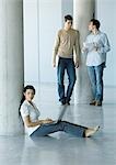 Young woman sitting on floor, using laptop, two men standing behind her