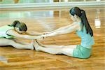 Two young women sitting on gym floor, feet to feet, stretching
