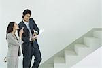 Man and woman in suits standing at foot of stairs, man looking at document, while woman points upwards