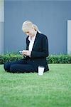Businesswoman sitting on grass with coffee, looking at cell phone, smiling