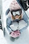 Preteen girl sitting on snow underneath propped up snowboards, high angle view