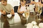 Boy and girl sitting at table, both holding Christmas ornaments, adults drinking champagne in background