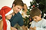 Father and two children opening Christmas present in front of Christmas tree, girl wearing Santa hat