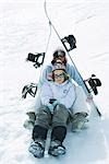 Two young skiers sitting under skis together, smiling at camera, portrait