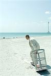 On beach, man in suit pulling garbage can towards ocean, side view, full length
