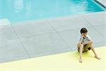 Boy sitting on the ground next to swimming pool, using cell phone
