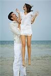 On beach, man holding young female companion up in air, full length
