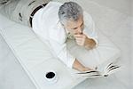 Mature homme couché sur le ventre sur une chaise longue, livre de lecture, tasse de café à proximité, vue grand angle