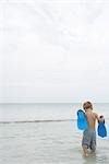 Little boy walking in shallow water, holding up flippers, rear view