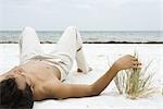 Young man lying on back on sand, touching dune grass, ocean in background