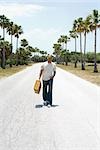 Man walking down center of road, carrying suitcase, listening to headphones