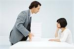 Businessman sliding tall stack of paper across a table to woman leaning on her elbow