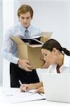 Femme écrivant sur le document au bureau d'homme portant une boîte en carton pleine de fournitures de bureau
