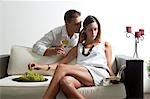 Young man leaning on couch with white wine whispering in young woman's ear reading book and eating grapes