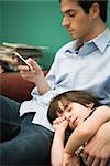 Young man text messaging while son naps with head in his lap