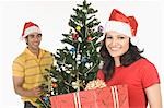 Woman holding a Christmas present with a man decorating a Christmas tree in the background