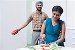 Woman chopping vegetables in the kitchen with her husband standing behind her with a present