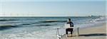 Man working at desk placed at water's edge on beach, wind turbines on horizon