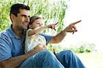 Father holding young daughter on lap outdoors, both pointing and looking away