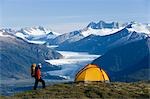 Hiker next to tent on ridge views Mendenhall Glacier & Coast Mountains near Juneau Alaska during Summer