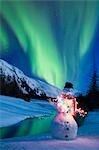 Lighted snowman decoration standing on riverbanks with northern lights display overhead Composite