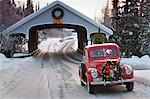 Man driving a vintage 1941 Ford pickup through a covered bridge with a Christmas wreath on the grill and a tree in the back during Winter in Southcentral, Alaska