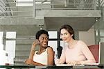 Two businesswomen in office, one using mobile phone, portrait