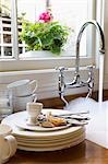 Stack of Dirty Dishes and silverware by Sink with running water below window with flower