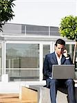 Businessman sitting alone in plaza using laptop and cell phone