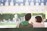 Young couple sitting on bench, looking at map, in park, back view