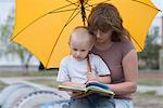 Young boy and woman sit under yellow umbrella reading a book