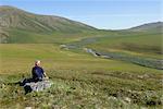 Senior male hiker resting on a rock outcrop enjoying the scenic view of tundra at Caribou Pass, ANWR, Arctic Alaska, Summer