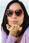 Woman wearing heart shaped sunglasses, blowing a kiss at camera