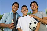 Boy (13-15) holding soccer ball with two brothers outdoors, front low angle view.