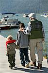 Grandfather and two grandsons holding fishing rods, walking on pier, back view