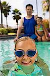 Girl (7-9) in swimming pool, father and sister (7-9) sitting on pool edge in background, front view portrait.