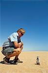 Africa, Namibia, Skeleton Coast. A meerkat (Suricata suricatta) stands typically alert as a local guide kneels close by.