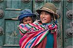 Peru, peruanische Mädchen trägt ihre kleine Schwester auf dem Rücken neben massiven Türen Kirchen Iglesia De la Compania de Jesus in Cusco s Plaza de Armas.